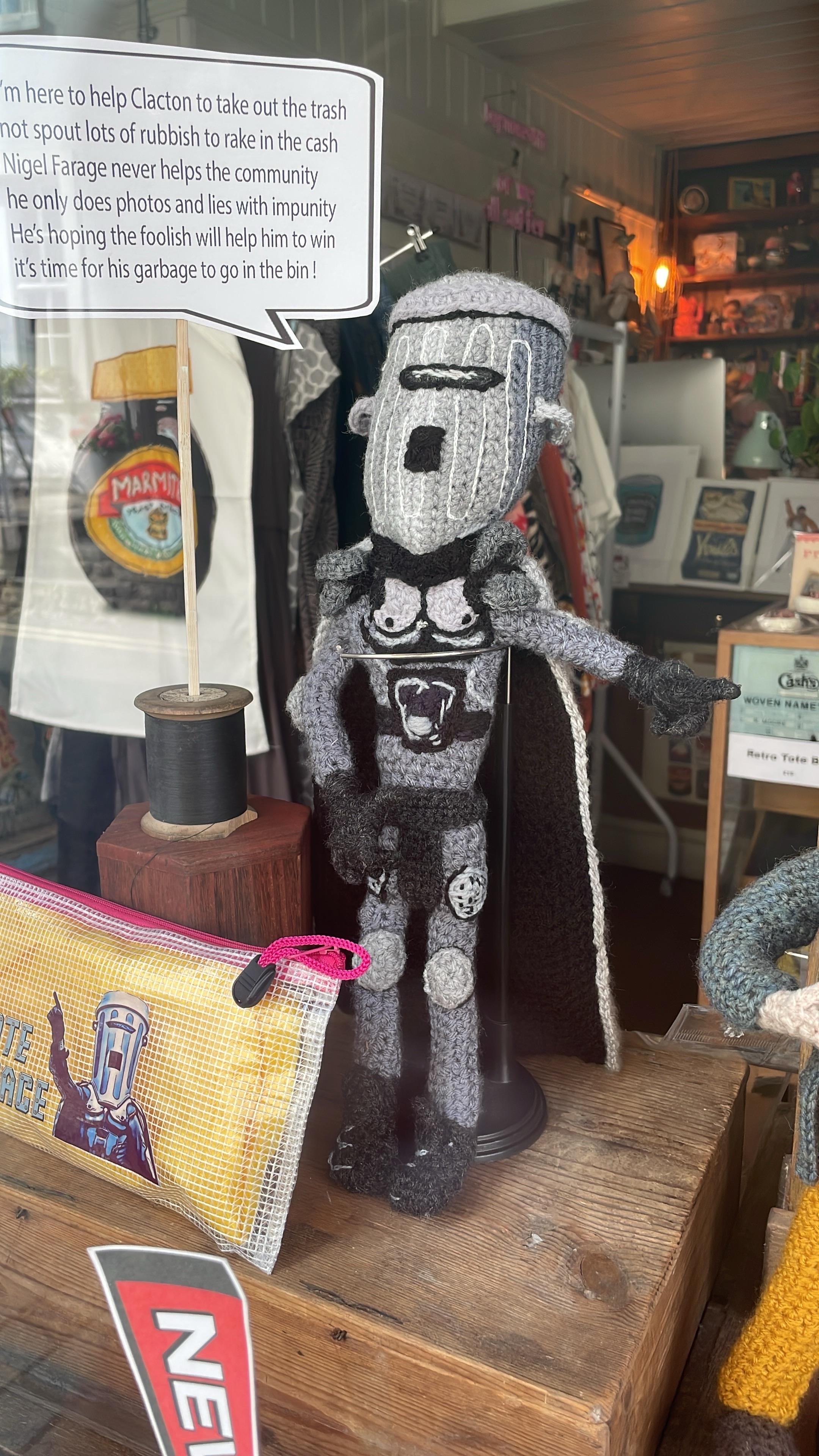 A crocheted and needle-felted figure, about 35cm high, in grey and black with a distinctive large space helmet and cape stands in a shop window. It is British national treasure, political saviour and all round hero COUNT BINFACE! A speech bubble coming out of his mouth reads: “I’m here to help Clacton to take out the trash / not spout lots of rubbish to rake in the cash / Nigel Farage never helps the community / he only does photos and lies with impunity / He’s hoping the foolish will help him to win / it’s time for his garbage to go in the bin!”